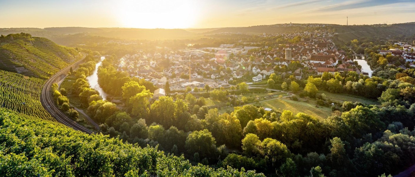 Das Bild zeigt den Blick von der Weinkanzel in Besigheim im Herbst. Das Motiv wird dominiert von Wein, der an den Steillagen angebaut wird. Im Tal ist der an dieser Stelle U-förmig verlaufende Neckar zu sehen. Im Hintergrund erkennt man die braunen Dächer der Stadt. Dieses Motiv nimmt die unteren 2/3 des Bilds ein. Darüber erstreckt sich blauer Himmel mit der tief stehenden Sonne., © Stuttgart-Marketing GmbH, Sarah Schmid